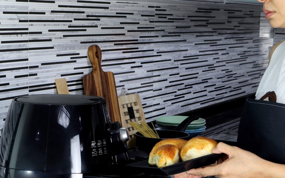 A mature woman in an apron sliding a drawer with pieces of bread on it into the tray of a black air fryer on her counter.
