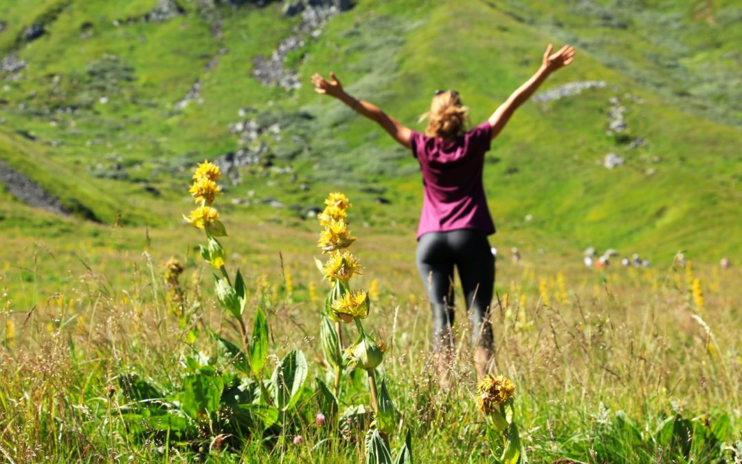 A happy woman in a t-shirt and leggings stands in a field with yellow flowers, with her arms raised, facing the mountains.