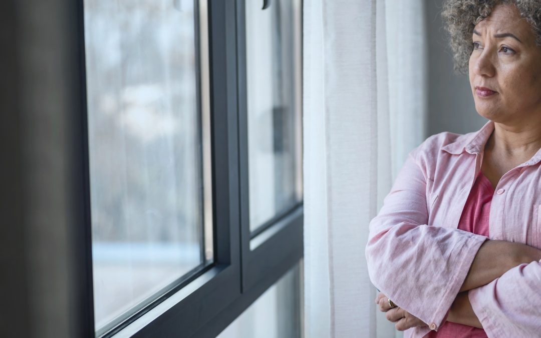 A middle-aged woman has her arms crossed and stares out of a window from a gray-walled room. She appears deep in thought.